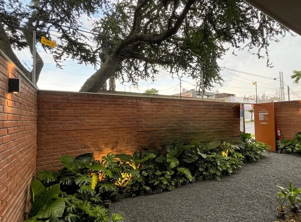 Jardín de llegada del departamento muestra: muro de ladrillo, follaje y el árbol que corona el acceso